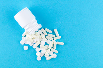 Close up white pill bottle with spilled out pills and capsules on blue background with copy space. Focus on foreground, soft bokeh. Pharmacy drugstore concept. Top view