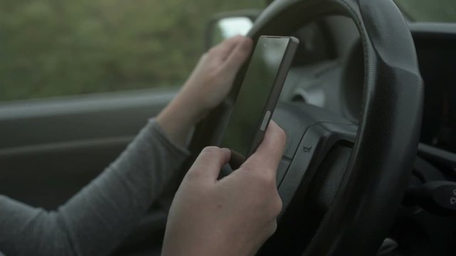 Woman simultaneously driving car and reading text message, mobile smart phone in female hands over vehicle steering wheel