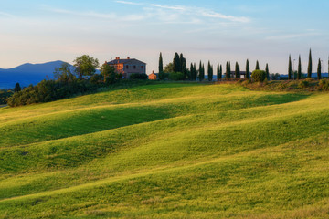 Fototapeta premium Magnificent spring landscape at sunrise.Beautiful view of typical tuscan farm house, green wave hills, cypresses trees, hay bales, olive trees, beautiful golden fields and meadows.Italy, Europe
