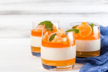 Creamy panna cotta with orange jelly in beautiful glasses, fresh ripe mandarin, blue textile on white wooden background. Delicious Italian dessert. Closeup photography.Selective focus.