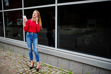 Portrait of a beautiful woman in red blouse and casual jeans talking on mobile phone and holding a cup of coffee outside the huge shopping mall.