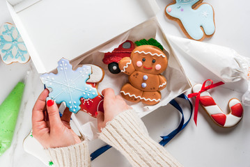 Obraz premium Preparing for Christmas, decorating traditional gingerbread with multicolored sugar glaze, the girl folds the cookies in a white gift box, with ribbon bow, on a white marble table copy space top view