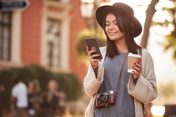 Pretty asian girl in modern wear using mobile phone and holding coffee to-go while walking on the city street