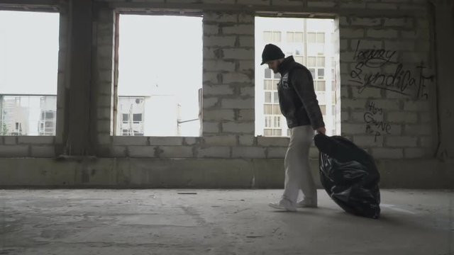 Homeless man hardly drags a garbage bag in abandoned building