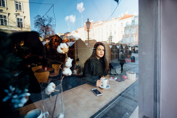 Beautiful brunette young woman in a cafe holding a cup of coffee or cocoa, seen through the window with buildings and lights reflections. She is looking away. Lifestyle concept.