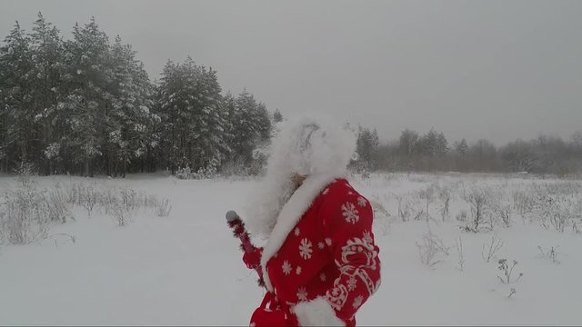 A merry Santa Claus with a bag of gifts walks in the winter snow-covered forest. New Year's and Christmas. Slow Motion Picture