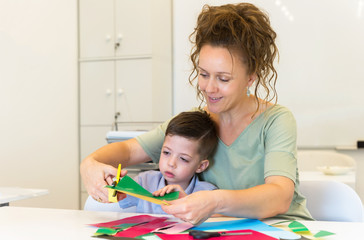 teacher woman and child boy cutting collage color paper with scissors in the classroom