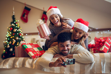 Fototapeta na wymiar Three playful positive modern friends lying on the bed together and taking a selfie with Santa hats and gifts for Christmas holidays.