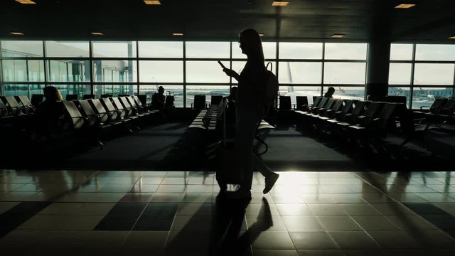 A woman is walking along the terminal of the airport, carrying a bag on wheels. In the hand smartphone