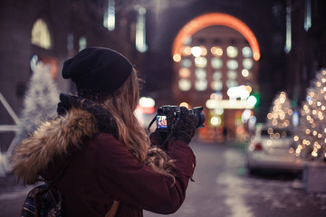 Traveler girl walking through the night city street taking photos of buildings and Christmas trees