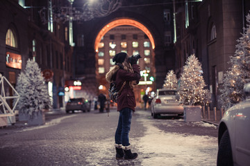 Obraz na płótnie Canvas Hipster girl walking through the night city street taking photos of buildings and Christmas trees