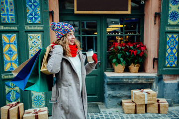 Attractive cute girl in with the shopping bags and holding disposable cup with warm drink in her hand. Female dressed in warm bright hat, red scarf and mittens. Happy holiday and freedom concept.