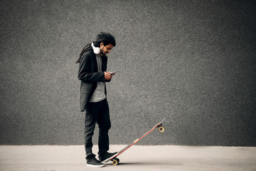 Stylish young dreadlocks hipster skater with headphones looking at smart phone standing on long board. © Dusan Petkovic