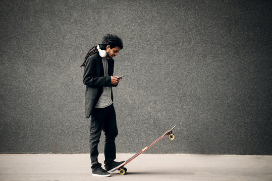 Stylish young dreadlocks hipster skater with headphones looking at smart phone standing on long board.