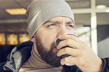 Adult bearded man in a beanie and dark jacket drinks hot coffee in a cafe at gas station, close up