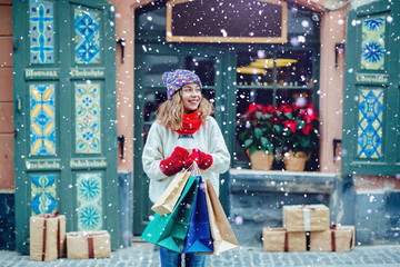 Obraz na płótnie Canvas Attractive girl with shopping bags holding disposable cup with warm drink in her hand.Woman near the shop with gifts enjoying winter and dressed in warm bright hat, red scarf and mittens.