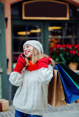 Beautiful smiling woman with shopping bags near shop with gift calling smart phone.