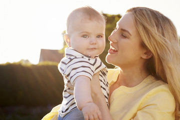 Young Mother Carrying Baby Son Outdoors In Summer Garden