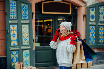 Beautiful smiling woman with shopping bags near shop with gift calling smart phone. Girl dressed in knitted hat, red scarf and mittens. Vacation, weekend, takeaway drinks, leisure concept