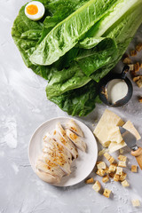 Ingredients for cooking classic Caesar salad. Sliced baked chicken breast, green roman salad, parmesan cheese, boiled egg, croutons, salt, jug of sauce over gray texture background. Top view, space.