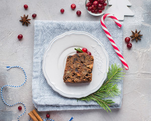 A piece of traditional Christmas English pudding (fruit cake) on white plate on a concrete light background. Christmas decor of the table. Funny napkins with reindeers Santa Claus.  Flat lay, top view
