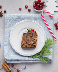 A piece of traditional Christmas English pudding (fruit cake) on white plate on a concrete light background. Christmas decor of the table. Funny napkins with reindeers Santa Claus.  Flat lay, top view