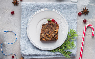 A piece of traditional Christmas English pudding (fruit cake) on white plate on a concrete light background. Christmas decor of the table. Funny napkins with reindeers Santa Claus.  Flat lay, top view
