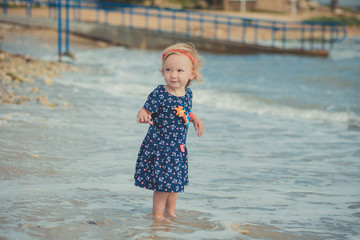 Baby cute girl with blond hair and pink apple cheek enjoying summer time holiday posing in beautiful beach full of sand wearing stylish dress