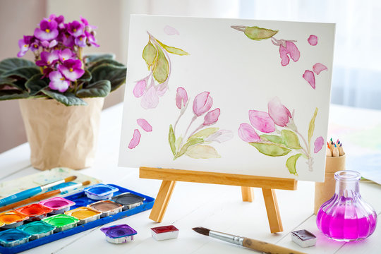 Canvas on easel, watercolor sketch, paints, brushes and violet senpolia flower in pot.
