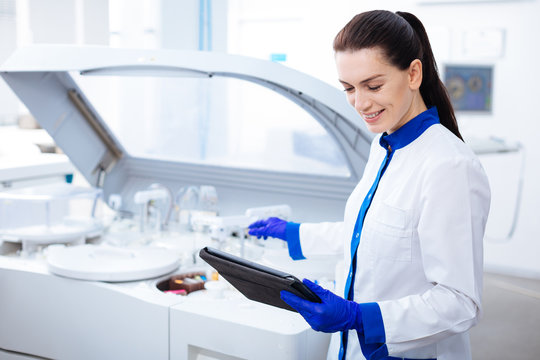 Science is light. Beautiful gentle inspired  female intern smiling adorably and looking at the tablet while stretching her hand to the centrifuge machine