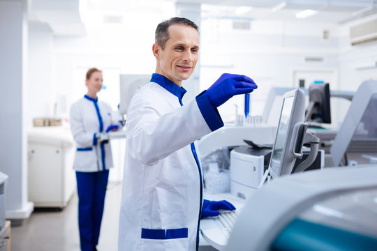 Let me see. Optimistic striking male researcher  smiling and typing while holding test glass in  a hand  near the medical equipment