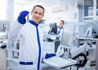 Successful experiment. Happy pleased male researcher achieving necessary reaction while placing his hand on the keyboard and brining up  glassware while standing in the lab 