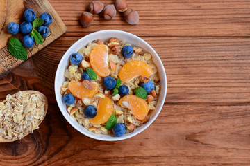 Oats porridge without sugar. Porridge with fresh tangerines, blueberries and hazelnuts in a white bowl and on a wooden background with copy space. What to put in porridge healthy. Top view