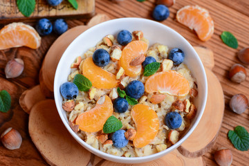 Oats porridge with berries and nuts. Homemade porridge with fresh tangerines, blueberries, hazelnuts and mint in a white bowl and on a wooden table. Simple way to make porridge interesting. Closeup