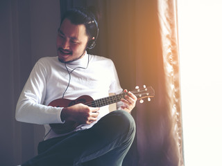 Obraz premium Young man in white t-shirt sitting in the room playing ukulele with earphone. Man practicing ukulele, having fun along.