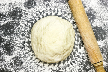 An overhead photo of wheat dough, rolling pin and sprinkled flour like a mandala on the dark background. Selective soft focus. Dough ready for cooking. Round circle shape on the flour.
