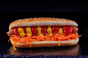 Fried chicken sausage, hot dog, with Korean carrots, ketchup and sweet mustard on a stony black background with reflection