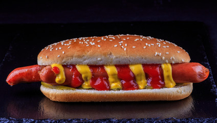 Spicy fried sausages in a bun with sesame, with mustard, ketchup, pickles and cheese on a black background