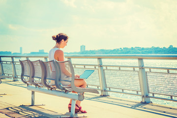 American woman traveling, working in New York.  A college student studying on summer vacation, sitting on chair at park by Hudson River, facing New Jersey, reading at laptop computer. Back View..