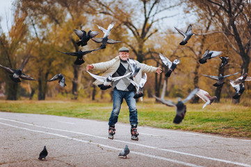 An elderly man has fun skating on roller skates. The man is spreading hands. People and pigeons.