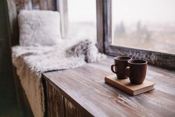 Brown cups stand on a book on wooden windowsill with blanket and pillow