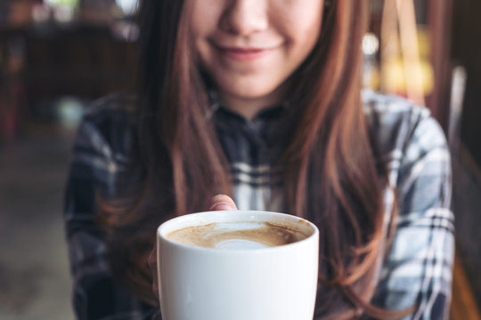 Closeup image of a beautiful Asian woman holding and showing a white mug while drinking hot coffee with feeling good in cafe