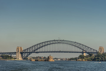 Fototapeta na wymiar Sydney, Australia - March 26, 2017: Frontal view of black metalic Harbour Bridge including support towers on both sided seen off water under blue sky. Denison Fort in the bay and Kirribilli high 