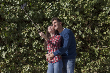 happy young couple taking a selfie with smartphone on the stick over green ivy background. The woman is holding the stick and shooting looking at phone with happiness. technology and love concept.