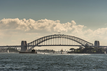 Fototapeta na wymiar Sydney, Australia - March 26, 2017: Frontal view of black metalic Harbour Bridge including support towers on both sided seen off water under cloudscape. Denison Fort in bay and multiple small boats.