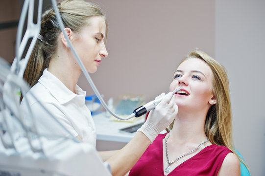 Attractive patient in red-violet dress laying on the dental chair while female dentist treating her teeth with special instruments.