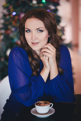 Beautiful mystic lady dressed in modish casual blue dress with brunette hairs and pout sensual lips sitting posing lonely in restaurant waiting her boyfriend husband with christmas tree on background