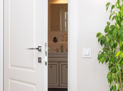 Ajar white door in a dark bathroom. Series switch on a light gray wall. Modern chrome door handle and lock