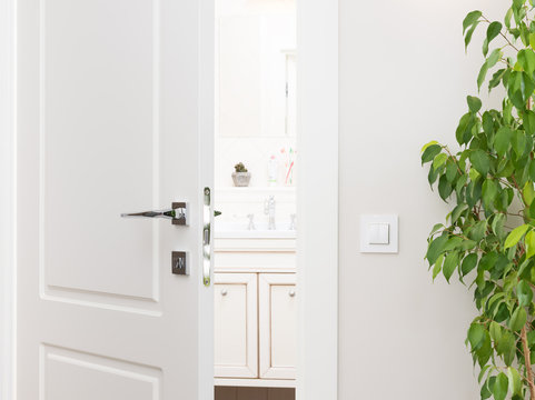 Ajar white door to the bathroom. Series switch on a light gray wall. Modern chrome door handle and lock