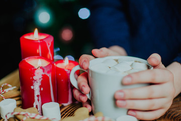 girl woman is holding a cup of cocoa with marshmallow on wooden background. With candles, ginger cookies, on christmas tree background 